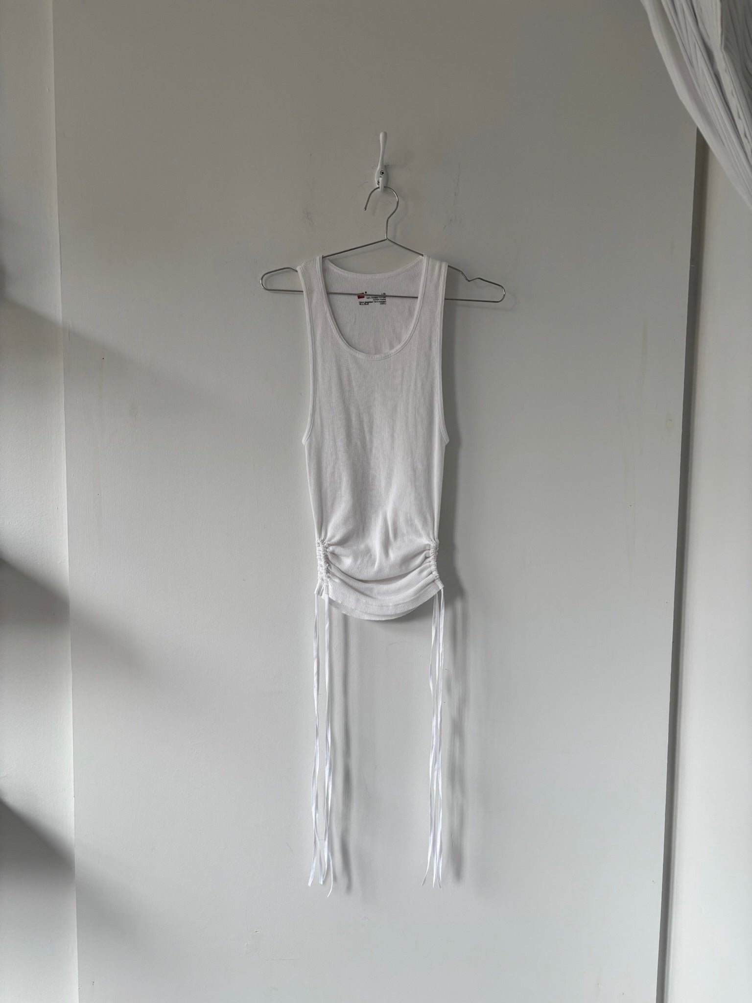 White tank top with fringe details hanging on a hook against a white wall.
